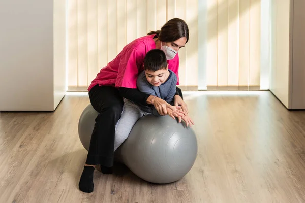 Niño discapacitado y fisioterapeuta en la parte superior de una pelota de gimnasio de cacahuete haciendo ejercicios de equilibrio. protección máscara pandémica —  Fotos de Stock