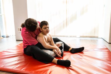 Kırmızı bir jimnastik minderinde egzersiz yapan engelli bir çocuk ve fizyoterapist. salgın maskesi koruması