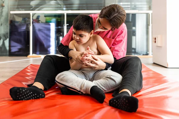 Mozgássérült gyermek és fizioterapeuta egy piros tornatermi szőnyegen gyakorlatokat végez. pandémiás maszkvédelem — Stock Fotó