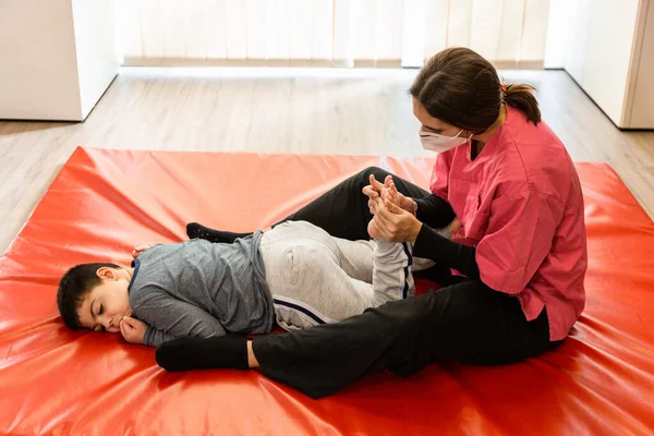 Niño discapacitado y fisioterapeuta en una alfombra de gimnasia roja haciendo ejercicios. protección máscara pandémica —  Fotos de Stock