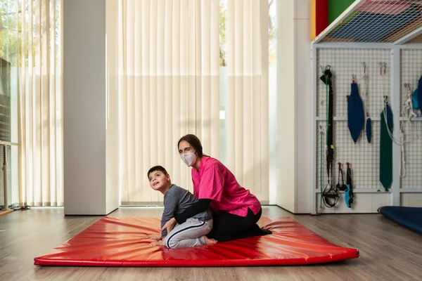 Niño discapacitado y fisioterapeuta en una alfombra de gimnasia roja haciendo ejercicios. protección máscara pandémica — Foto de Stock