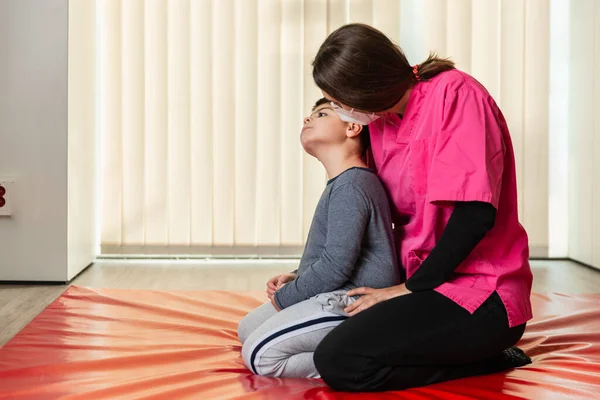 Kırmızı bir jimnastik minderinde egzersiz yapan engelli bir çocuk ve fizyoterapist. salgın maskesi koruması — Stok fotoğraf