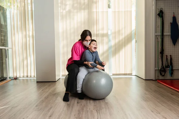 Mozgássérült gyermek és fizioterapeuta egy mogyoró tornaterem labda tetején, akik egyensúlyozó gyakorlatokat végeznek. pandémiás maszkvédelem — Stock Fotó