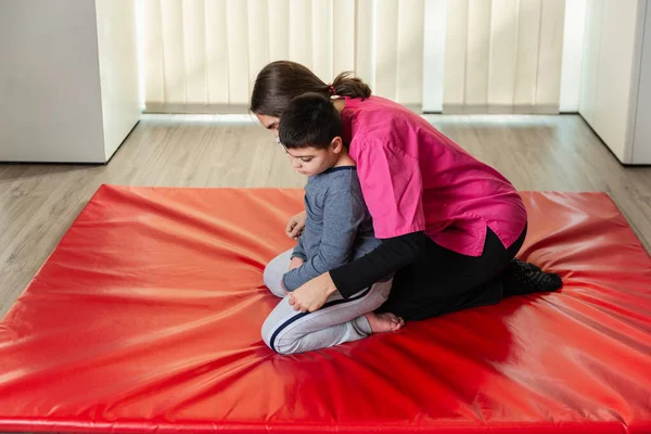 Niño discapacitado y fisioterapeuta en una alfombra de gimnasia roja haciendo ejercicios. protección máscara pandémica —  Fotos de Stock