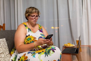 Kadın elinde cep telefonuyla kanepede oturuyor, dizüstü bilgisayarında yemek var.