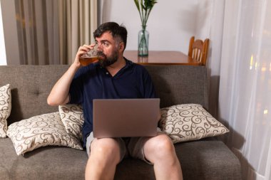 Kanepedeki sakallı adam dizüstü bilgisayar kullanıyor ve yemek odasında bira içiyor.