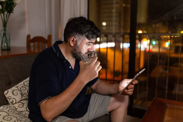 Kanepedeki sakallı adam dijital tablet kullanıyor ve yemek odasında bira içiyor. — Stok fotoğraf