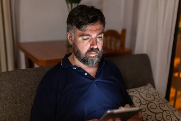 bearded man on sofa uses digital tablet in dining room overnight