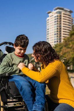 Sevgi dolu bir anne engelli oğlunu teselli ederken bir parkta tekerlekli sandalyede ellerini tutarak destek ve şefkat gösteriyor.