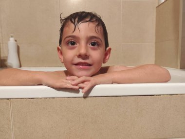 Islak saçlarıyla küvette gülümseyen, eğlenceli banyo yapan ve evde doğal ışık altında hijyen uygulayan genç bir çocuk.