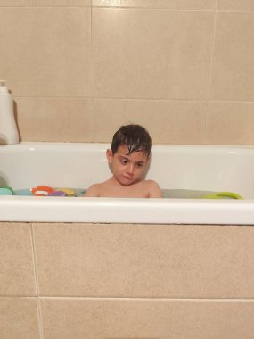 Islak çocuk banyoda renkli banyo oyuncaklarıyla çevriliyken küvette üzgün oturuyor.