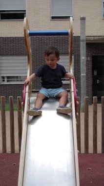 Açık hava parkında metal bir kayaktan kayan mutlu küçük çocuk. Bir çocuğun oyun parkı ekipmanlarıyla eğlendiği yavaş çekim videosu