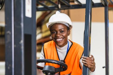 Afro-Amerikan kadın forklift kullanıyor, çelik yelek ve şapka giyiyor, modern bir depoda kameraya gülümsüyor.