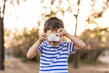 Elinde ahşap bir fotoğraf makinesi olan genç bir çocuk, altın saatinde açık hava parkında fotoğraf çekiyormuş gibi yapıyor, hayal gücüne ilham veriyor.