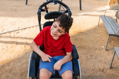 Serebral felçli ve zihinsel engelli bir çocuk tekerlekli sandalyesinde oturuyor, gülüyor ve açık hava parkında gülüyor.