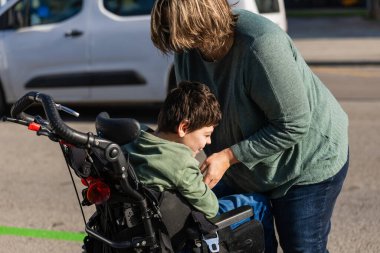 Mother placing her nine year old son with cerebral palsy into a wheelchair outdoors on a sunny day