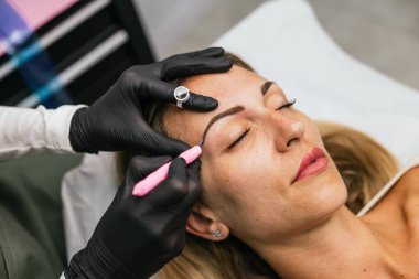 Kaşlarını geliştirmek ve şekillendirmek için yetenekli bir teknisyen tarafından mikro patinaj tedavisi gören bir kadın müşterinin yakından görüntüsü.