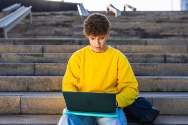 Üniversite kampüsünün dış basamaklarında oturan genç adam dizüstü bilgisayarla çalışıyor ve güneşli günün tadını çıkarırken derslere odaklanıyor.