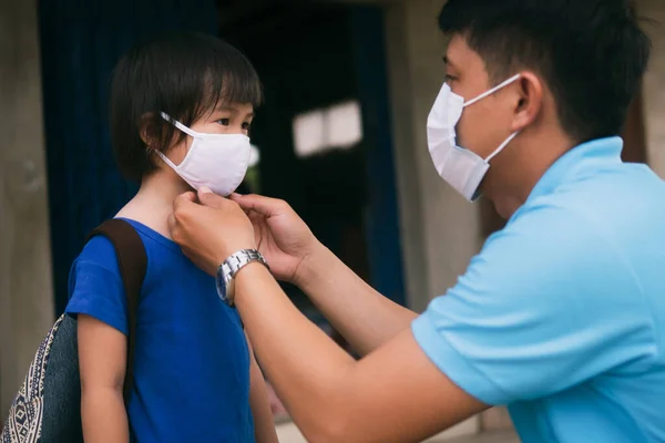 Asian father wearing protective mask puts protective cloth face mask on his little child, schoolkid with backpack is ready to school after Coronavirus COVID-19 pandemic.