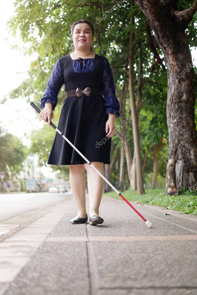 Asian blind person woman walking on sidewalk with a long white cane a mobility tool used to detect objects in the path, also helpful for onlookers in identifying the user as blind or vision disability