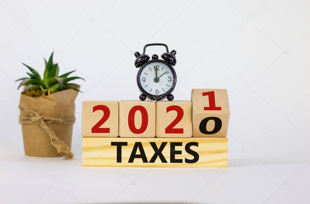 Business and new year concept of taxes planning 2021. Fliped wooden cube and changed words 'TAXES 2020' to 'TAXES 2021'. Black alarm clock and house plant. Beautiful white background, copy space.