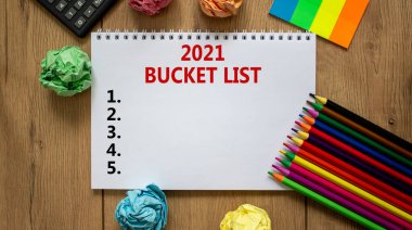 2021 bucket list symbol. White note with inscription '2021 bucket list' on beautiful wooden table, colored paper, colored pencils, paper clips, coins and calculator. Business and 2021 bucket list concept. clipart