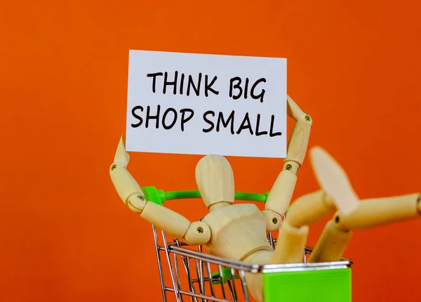 Think big shop small symbol. White sheet of paper. Words \'Think big shop small\'. Wooden model of a human in a shopping cart. Beautiful orange background. Business and sale concept, copy space.