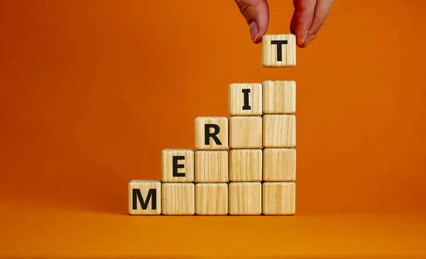 Merit symbol. Wood cubes with word \'merit\' stacking as step stair on beautiful orange background, copy space. Male hand. Business and merit concept.