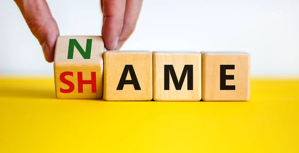 Name or shame symbol. Businessman turns the wooden cube and changes the word \'shame\' to \'name\' or vice versa. Beautiful yellow table, white background, copy space. Business, name or shame concept.