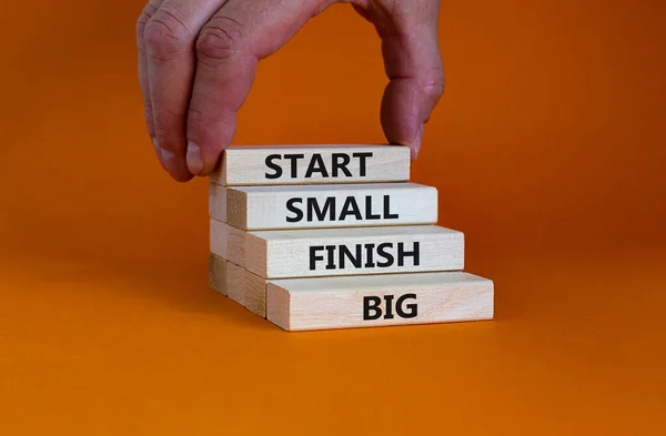 Start small finish big symbol. Concept words \'Start small finish big\' on wooden blocks on a beautiful orange background. Businessman hand. Business, motivational and start small finish big concept.