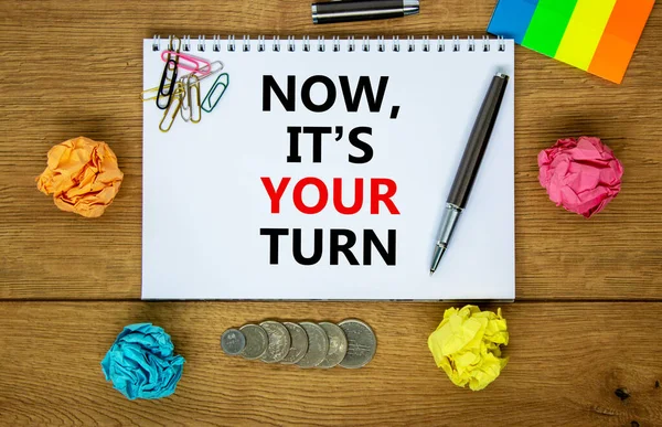 Now it is your turn symbol. Words \'Now it is your turn\' on white note. Wooden table, colored paper, paper clips, pen, coins. Business, Now it is your turn concept. Copy space.