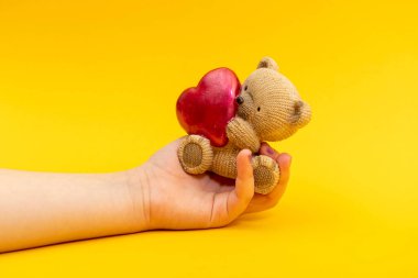 Genç bir kadın eli, sarı arka planda kırmızı kalpli küçük bir oyuncak ayıyı tutuyor. Sevgililer Günü konsepti.