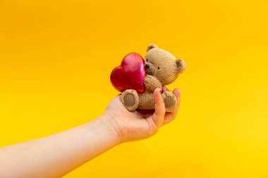 Genç bir kadın eli, sarı arka planda kırmızı kalpli küçük bir oyuncak ayıyı tutuyor. Sevgililer Günü konsepti.