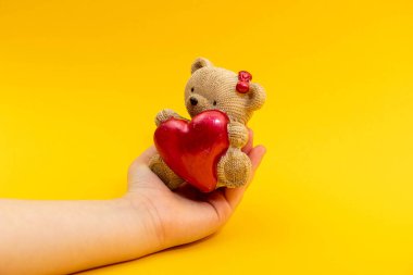 Genç bir kadın eli, sarı arka planda kırmızı kalpli küçük bir oyuncak ayıyı tutuyor. Sevgililer Günü konsepti.