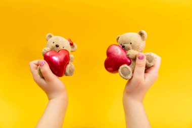 Genç bir kadın eli, sarı arka planda kırmızı kalpli küçük bir oyuncak ayıyı tutuyor. Sevgililer Günü konsepti.
