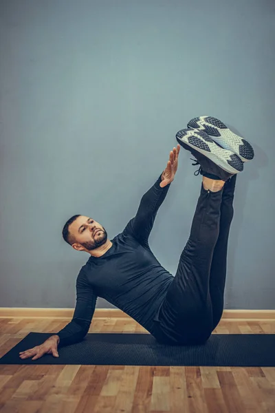 Confident young fitness man exercising on a fitness mat indoors, over gray wall background.