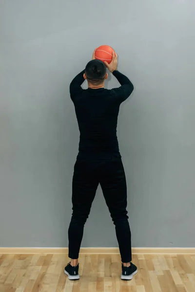 Young male athlete crouched doing wall balls exercises at the gym on grey background.
