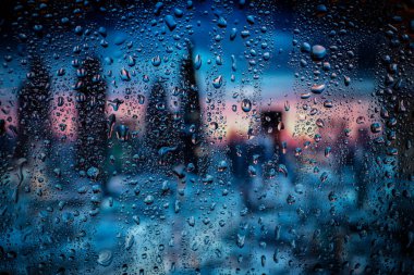 Pencerede yağmur damlaları, sırt çantaları şehrin ışıklarını bulanıklaştırıyor. Gece şehir hayatı yağmurlu mevsim soyut arka planında.