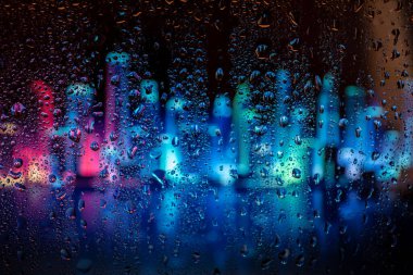 Pencerede yağmur damlaları, sırt çantaları şehrin ışıklarını bulanıklaştırıyor. Gece şehir hayatı yağmurlu mevsim soyut arka planında.