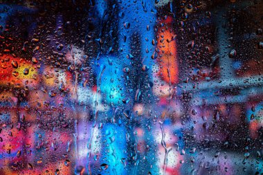 Pencerede yağmur damlaları, sırt çantaları şehrin ışıklarını bulanıklaştırıyor. Gece şehir hayatı yağmurlu mevsim soyut arka planında.