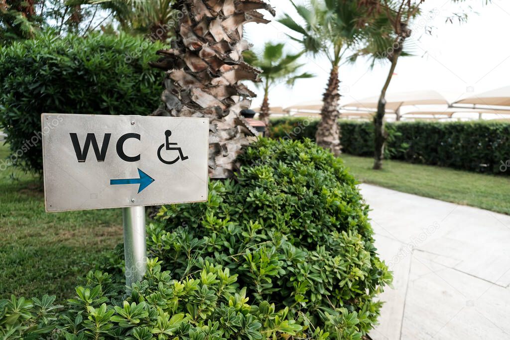 WC sign to a toilet room or washroom or bathroom or restroom including service for invalids or disabled people. Beautiful green tropical trees and bushes during summer. Board with a direction arrow.