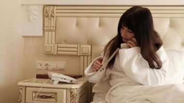 Beyaz otel bornozlu genç esmer kadın bir yatakta oturuyor ve telefonda bir arkadaşıyla konuşuyor, gülümsüyor. Otel odasındaki telefon konuşmaları. Beyaz kız tatilde bir arkadaşını arıyor..