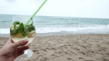 Female hand holding a glass with mohito cocktail at the beach. Refreshing beverage at a luxury getaway near the sea. Waves coming in the background. Plastic straw in a beverage with mint and lemon.