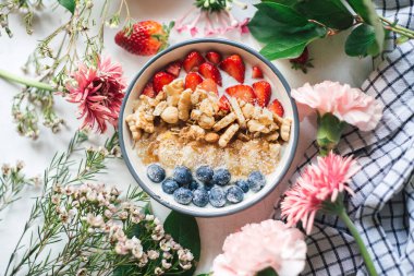 Son moda kahvaltı beşiği: yemyeşil fındık, tahıl, çilek, yabanmersini, muz dilimleri ve pembe çiçekli yoğurt. Taze meyveli sağlıklı müsli. Yemek tasarımı. Üst görünüm.