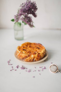 Beyaz masada taze pişmiş ricotta peynirli kek ve cam vazoda leylak çiçeği. Ev yapımı, organik malzemeli köy pastası. Peynirli ve dutlu unsuz turta. Yemek tasarımı, seçici odaklanma.