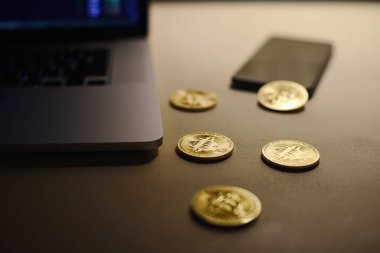Dizüstü bilgisayarı ve cep telefonu ticareti, seçici odak noktası ve tahıl dokusu olan bir masada altın bitcoin paralar