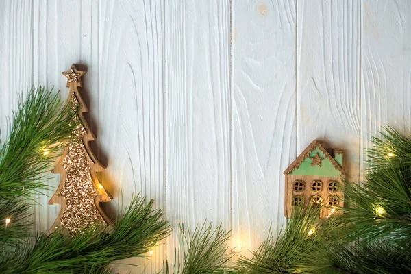 輝く窓と緑の松の枝と白い木製の背景に輝きを持つ輝く木製のクリスマスツリーを持つ小さな装飾的な木製の家 休日のために家にいるという概念 創作グリーティングカード ストックフォト