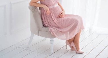 Açık pembe elbiseli ve pembe ayakkabılı genç hamile bir kız koltuğa oturur ve karnına dokunur.