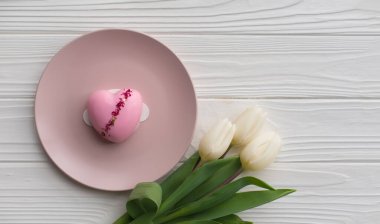 Beyaz ahşap arka planda bir buket beyaz lalenin yanındaki tabakta duran kalp şeklinde pembe bir pastanın üst görüntüsü. Sevgililer Günü 'nü kutlarım. Doğum günün kutlu olsun, Kadınlar Günü. Zarif tebrik kartı.