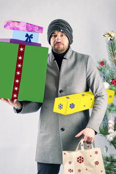 Noel alışverişi yapan adam. Kolları hediye kutularıyla dolu. Arka planda bir Noel ağacı var..